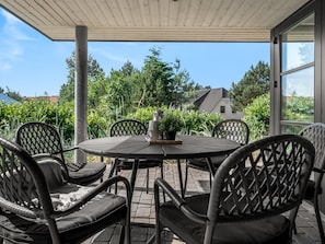 Une terrasse couverte avec une table ronde et six chaises, entourée de verdure et avec une maison en arrière-plan.