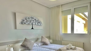 Habitación de hotel con dos camas, una mesita de noche con lámpara, una ventana con cortinas y un cuadro colgado en la pared con un árbol.