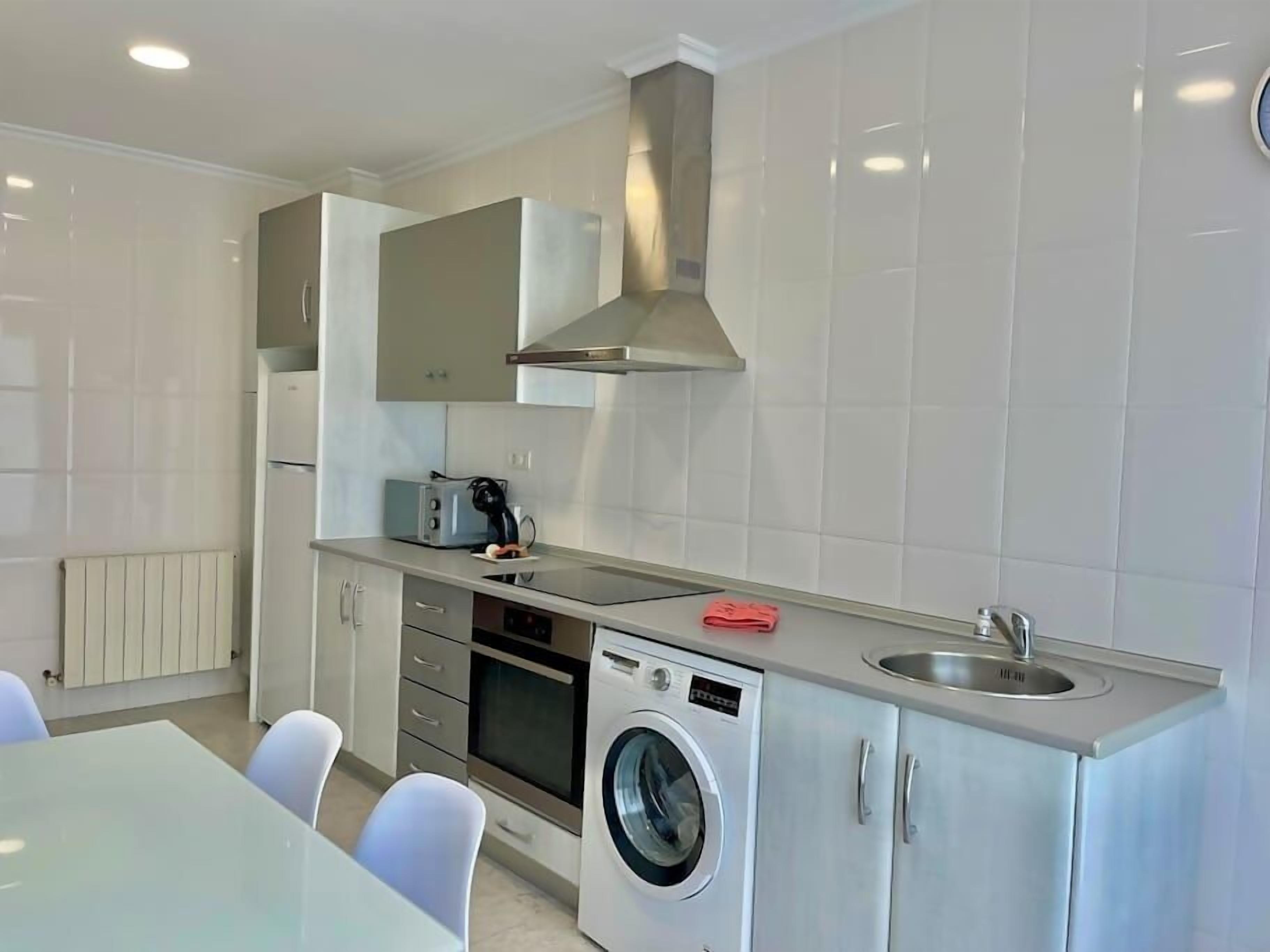 A modern kitchen with white cabinets, a built-in oven, a microwave, a washing machine, and a sink.