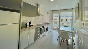 Una cocina moderna con horno empotrado, microondas y lavadora. Hay una zona de comedor con una mesa y sillas, y un ventanal con vistas a la ciudad.