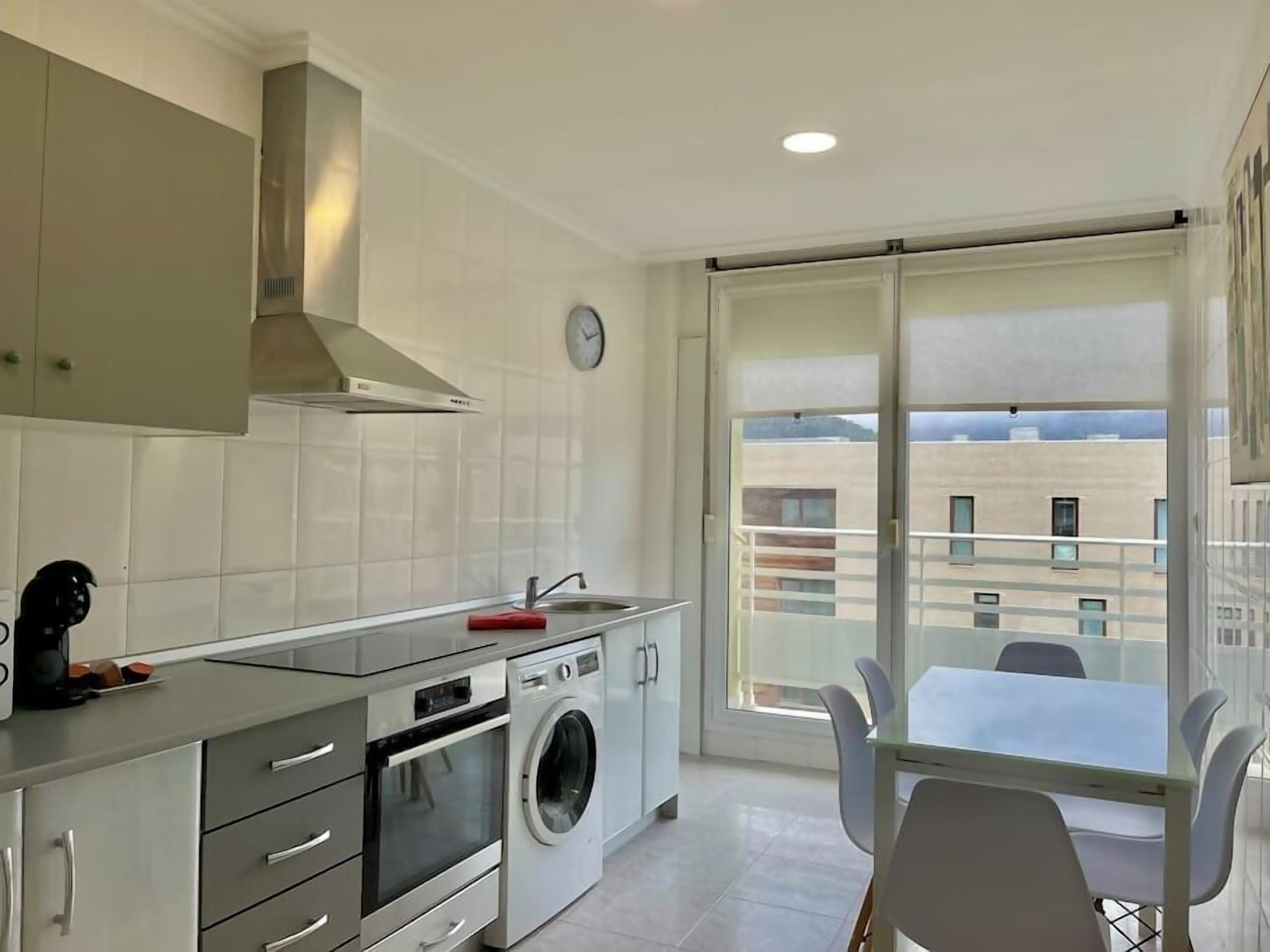 Cocina moderna con horno empotrado, microondas y lavadora. Cuenta con una zona de comedor con mesa y sillas, y un amplio ventanal con vistas a edificios.