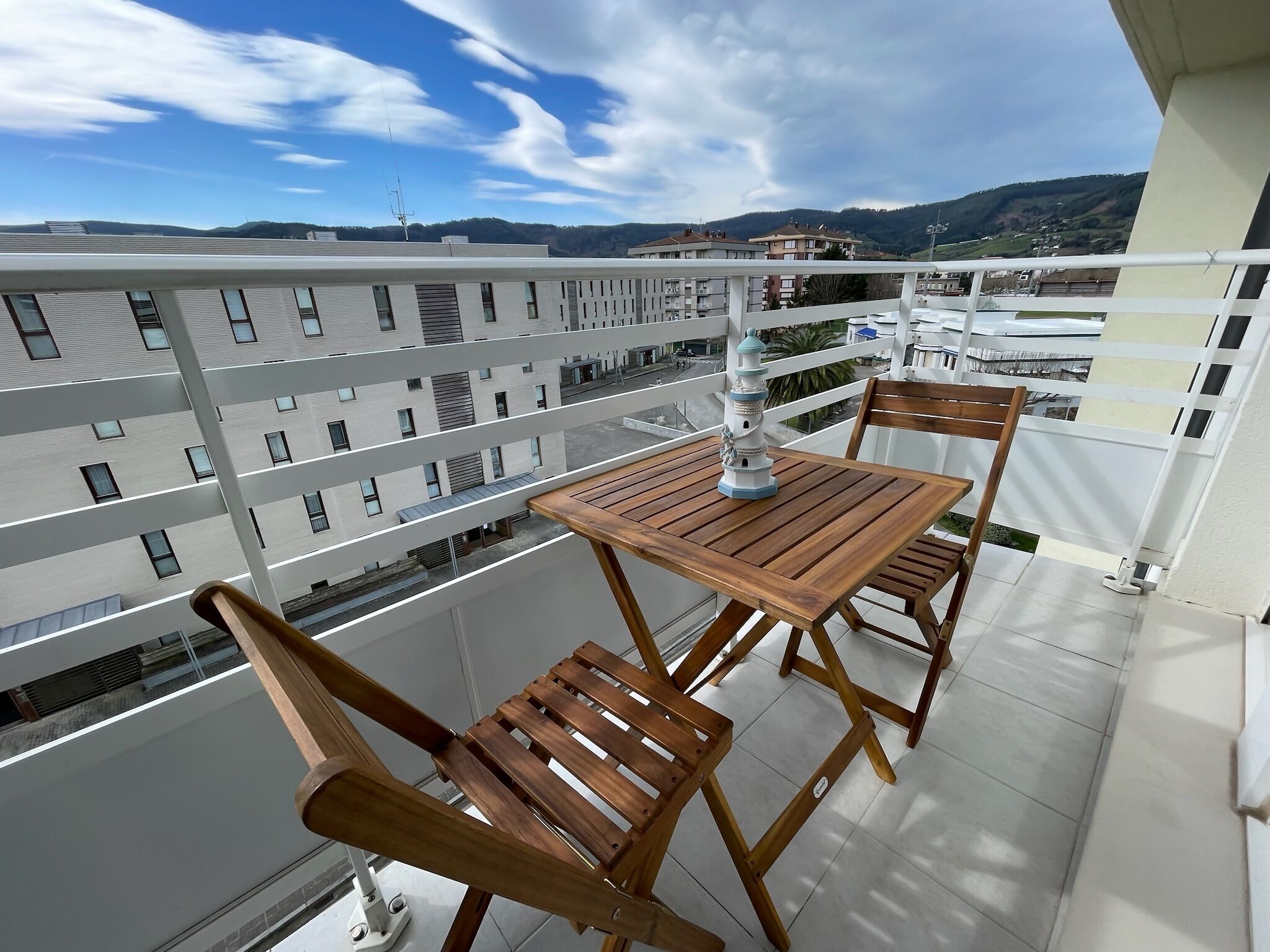 A balcony with a wooden table and chairs, a decorative windmill, and a view of a residential area with buildings and hills in the background.
