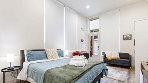 A modern hotel room with a large bed, a bedside table, a small desk, a sofa, and a view of the outside. - Perch - Cozy Studio | Own Bath | Heart of Fremont (Seattle)
