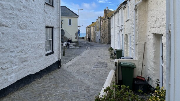 A narrow cobblestone street flanked by white buildings with stone facades, a green trash bin, and a broom leaning against a wall.