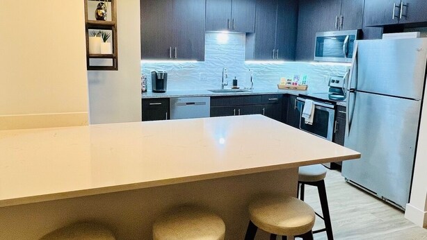 Une cuisine moderne dotée d’un grand îlot central, de tabourets de bar, de meubles de couleur sombre, d’appareils électroménagers en acier inoxydable et d’un luminaire suspendu.