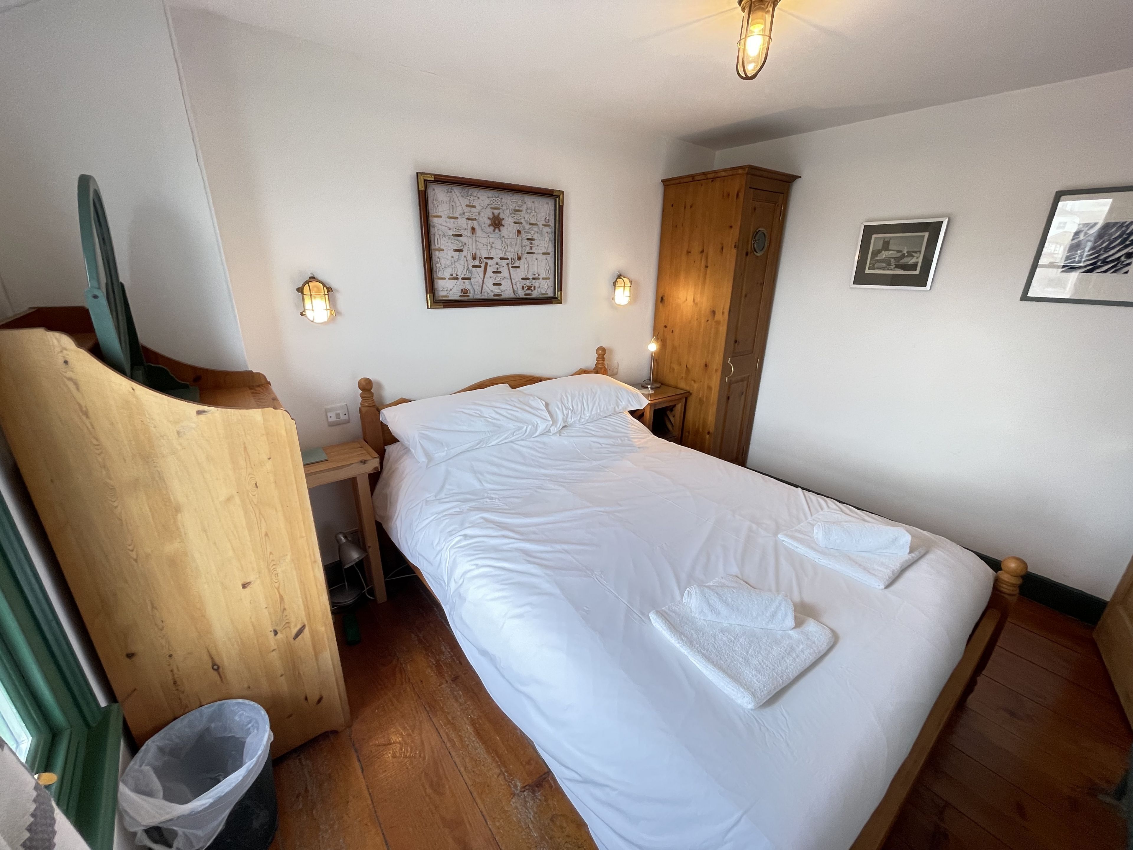 Een keurig opgemaakt bed met witte lakens en een opgevouwen handdoek, een houten garderobe, een ingelijst schilderij aan de muur en een klein nachtkastje met een lamp.