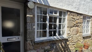 Un bâtiment en pierre avec une porte et des fenêtres blanches, un petit espace extérieur agrémenté de plantes en pot et une grille métallique fixée au mur.