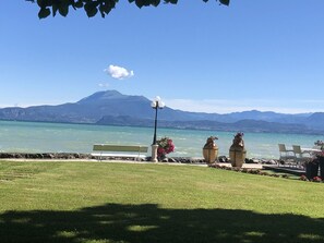 A grassy area with a bench, two large flower pots, and a table with chairs facing a body of water and mountains in the background. - Oasi dell'Anima By Bookinggardalake (Sirmione)