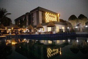 Un hôtel doté d’une piscine, d’un espace extérieur aménagé avec des parasols en chaume, et d’une façade moderne ornée de motifs lumineux.