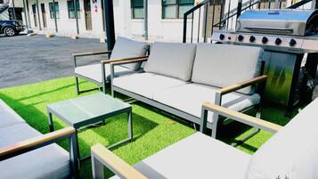 An outdoor seating area with a sofa, chairs, and a table, a barbecue grill, and a building in the background.