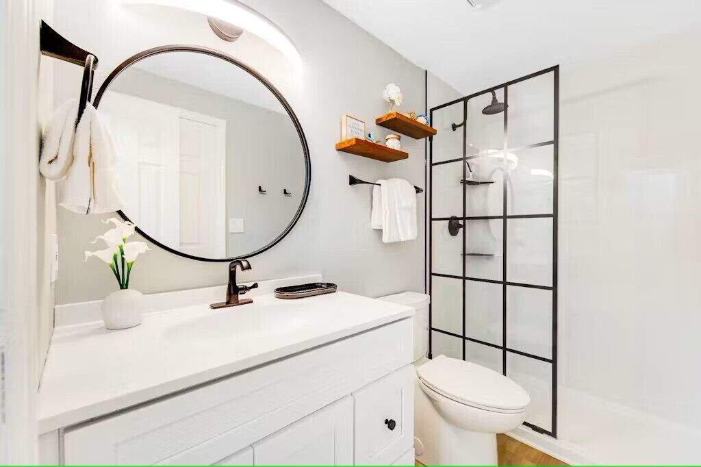 A modern bathroom with a white vanity, a round mirror, a toilet, and a glass shower enclosure.