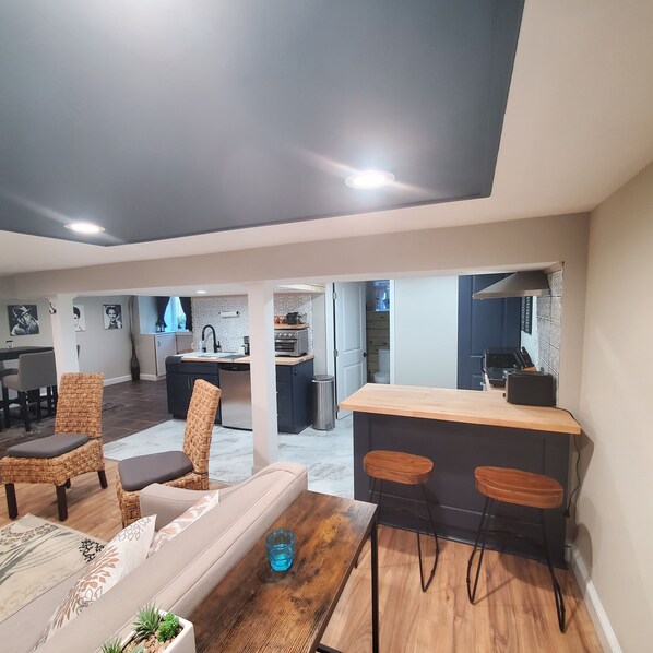 A modern living room with a sofa, two wicker chairs, a coffee table, and a kitchen area with a bar counter and stools.