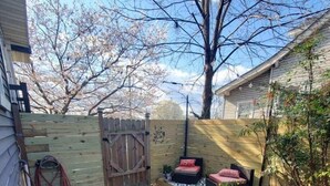 Un giardino recintato da una staccionata in legno, con un piccolo angolo relax con cuscini e un'area verde con diverse piante e un vialetto in ghiaia.