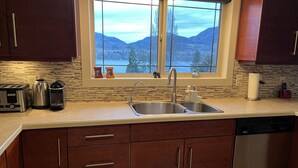 A kitchen with a stainless steel sink, a toaster, a kettle, and a coffee maker. There is a window with a view of mountains and trees. - Tranquil setting with beautiful views overlooking Skaha Lake  (Okanagan Falls)