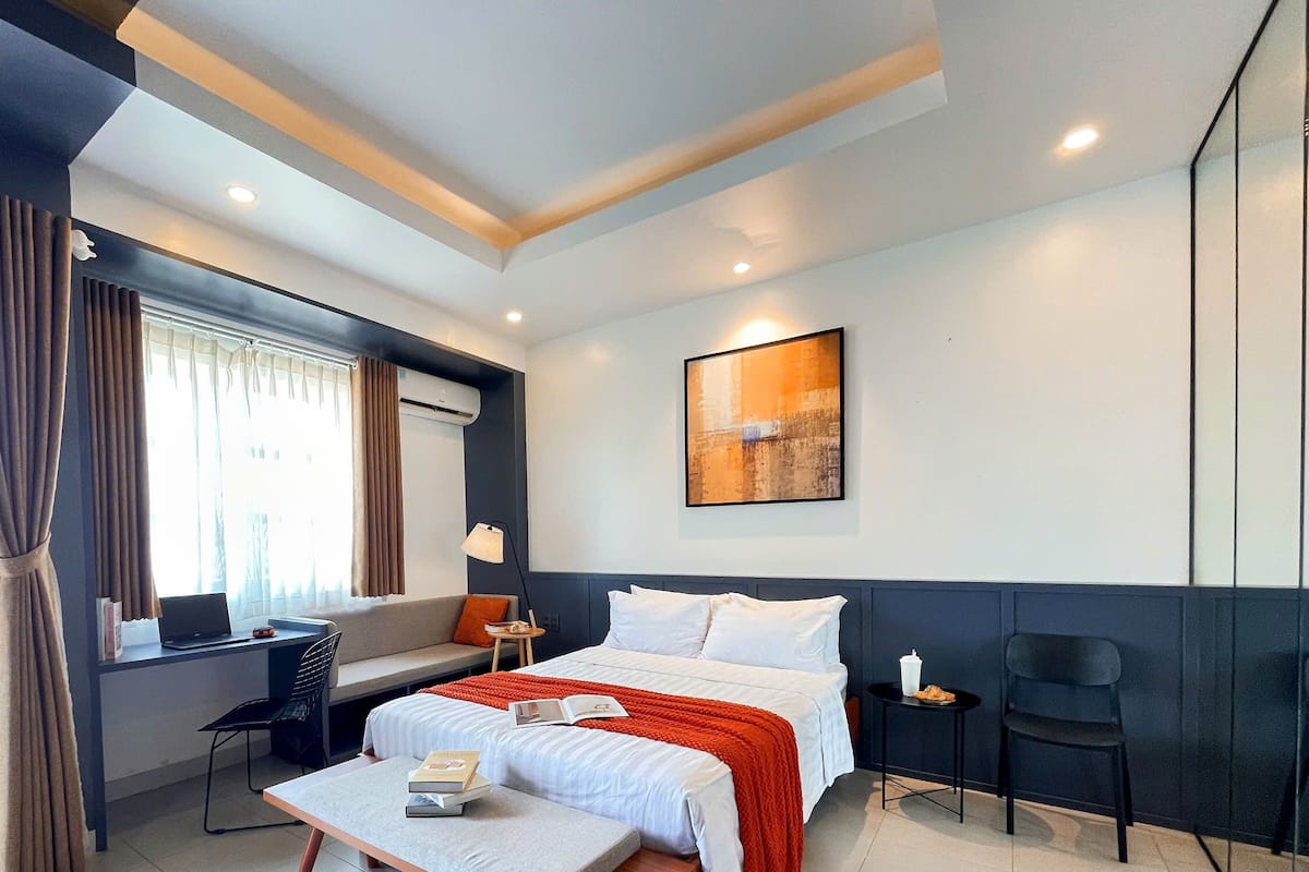 A modern hotel room with a bed, a desk, a chair, a small table, and a window with curtains.