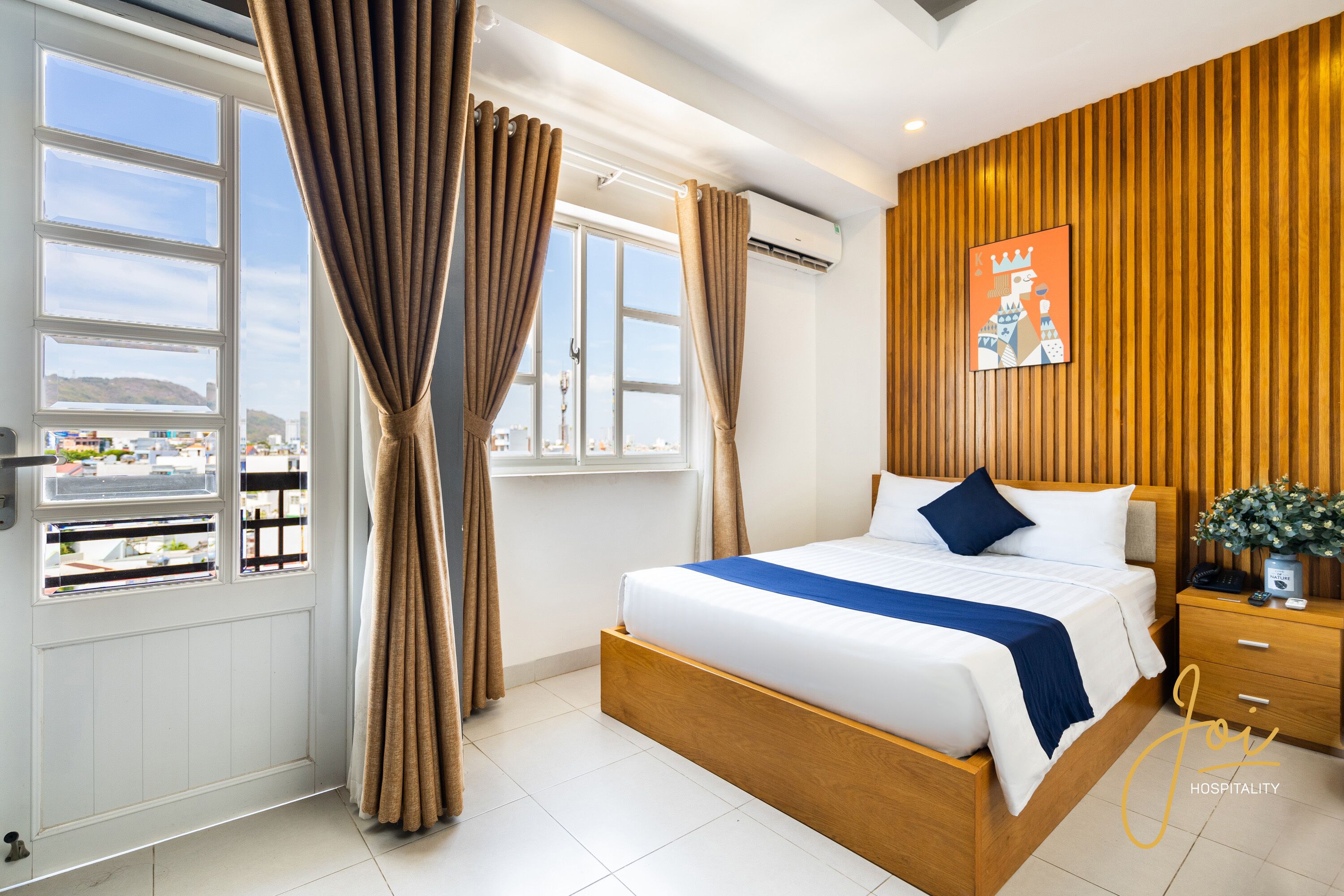 Una habitación de hotel moderna con una cama grande, cabecera de madera, mesita de noche y vista a la ciudad a través de amplios ventanales.