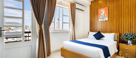 Una habitación de hotel moderna con una cama grande, cabecera de madera, mesita de noche y vista a la ciudad a través de amplios ventanales.