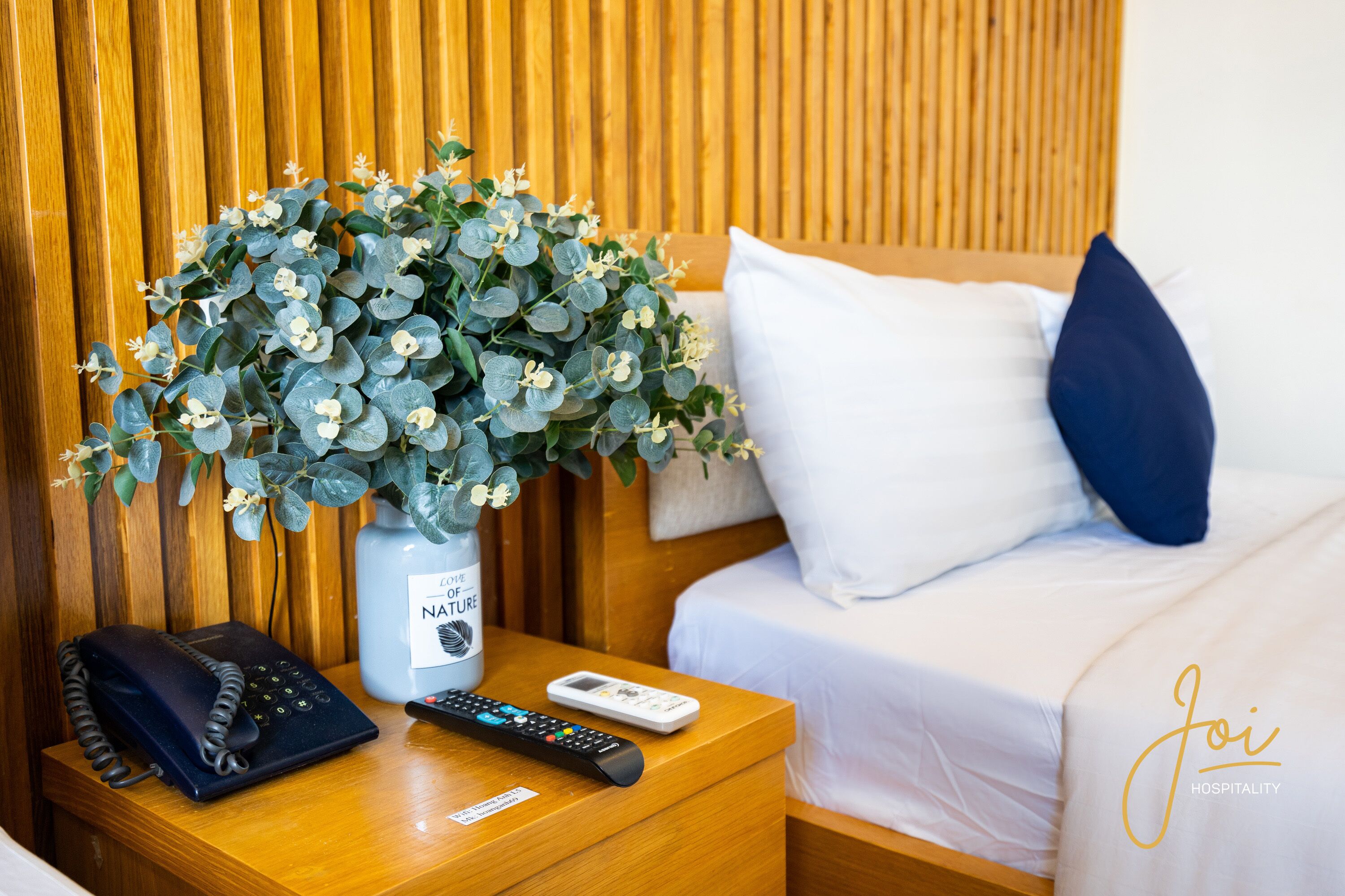 A neatly made bed with white and blue pillows, a bedside table with a telephone, a remote control, and a vase with a plant.