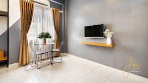 A modern living room with a flat-screen TV mounted on a grey wall, a small wooden shelf with a vase, a dining table with two chairs, and a potted plant. - Joi Hospitality - Hoang Anh (Ho Chi Minh City)