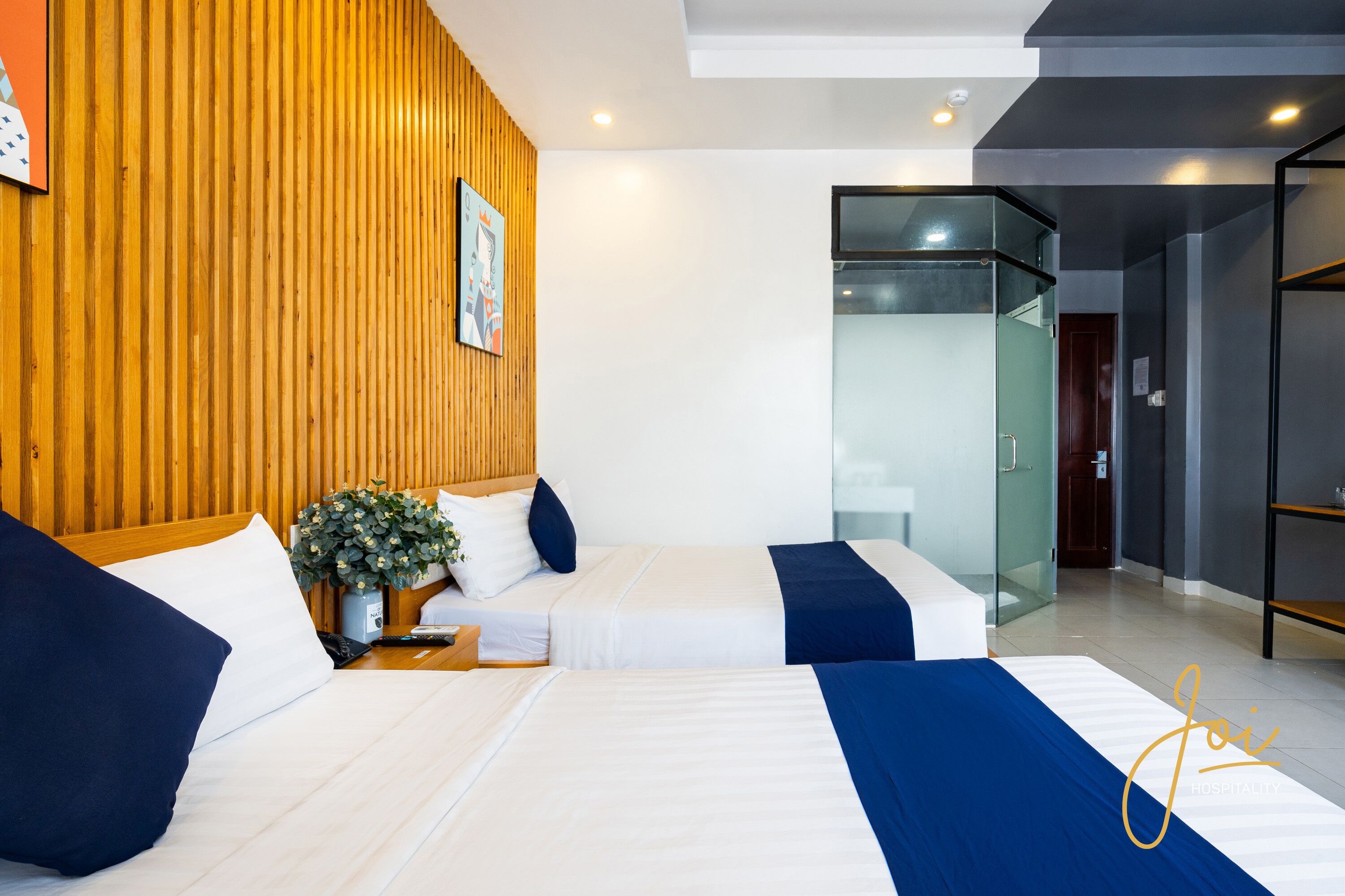 Hotelkamer met twee bedden, houten lambrisering aan de muur, een kleine plant en een glazen wand die de kamer van een ander deel scheidt.