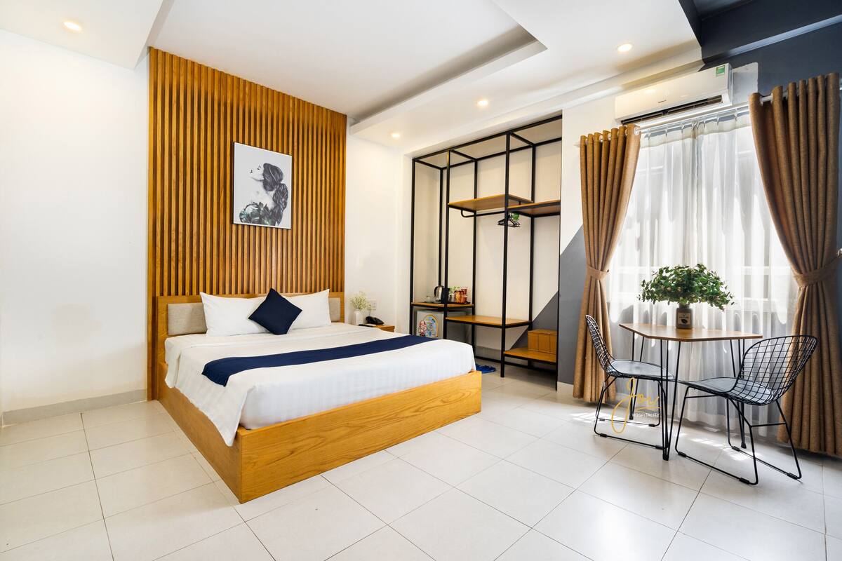 A modern hotel room with a wooden bed, a small table with chairs, a potted plant, and a view of the outdoors through sheer curtains.