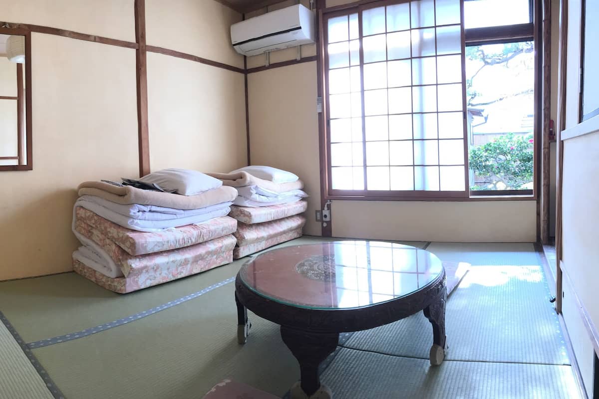 A traditional Japanese room with tatami flooring, a low table, a stack of futons, a wooden ladder, and a large window.