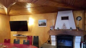 A dining area with a red table and chairs, a flat-screen TV mounted on the wall, a fireplace with a stone surround, and a clock on the wall. - Maison de Village (Viella)