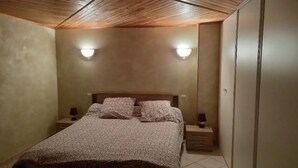 A bedroom with a wooden ceiling, a bed with floral bedding, two bedside lamps, and a wardrobe. - Maison de Village (Viella)