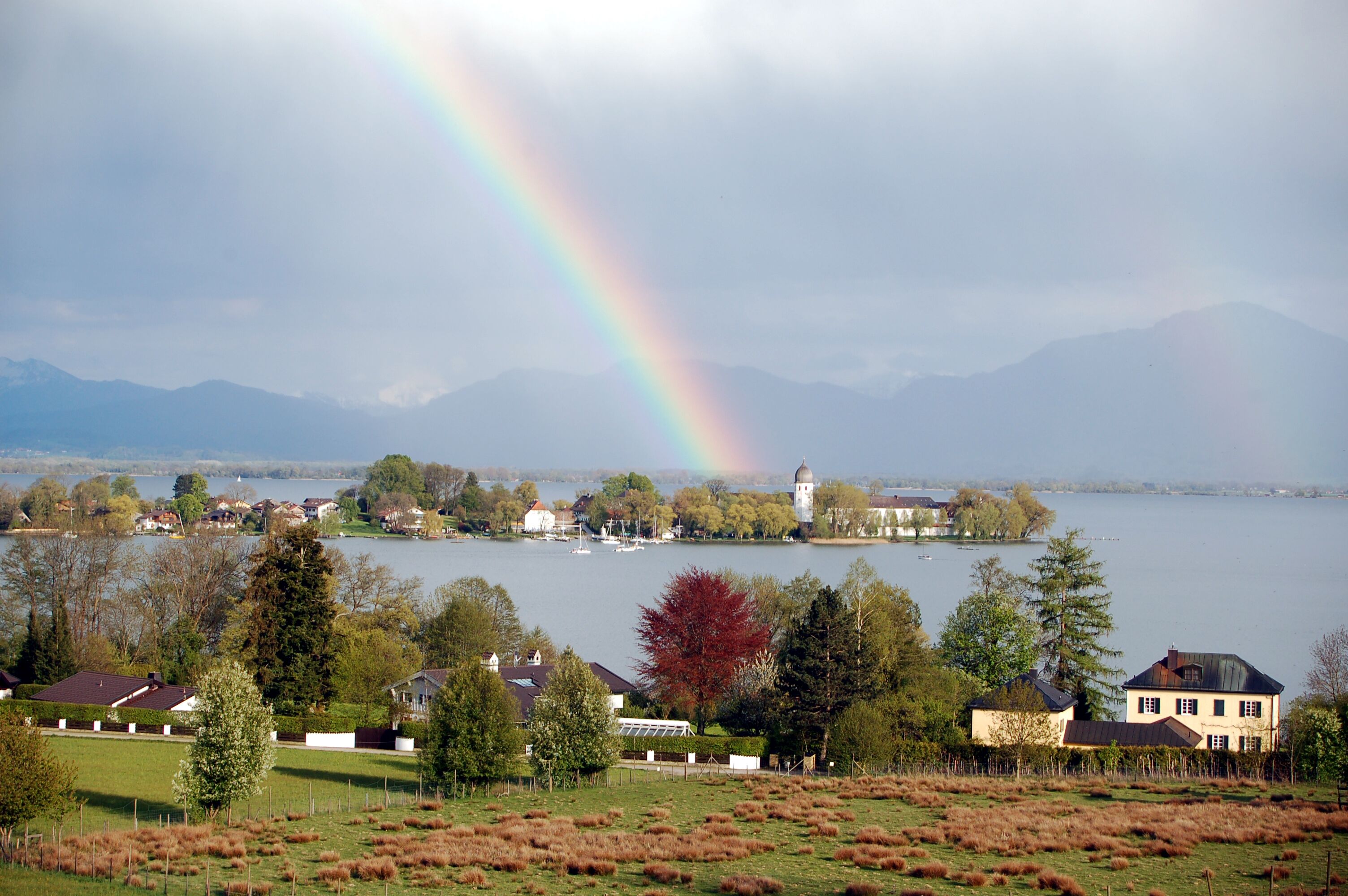A lakeside view with houses, a lighthouse, and mountains in the background.