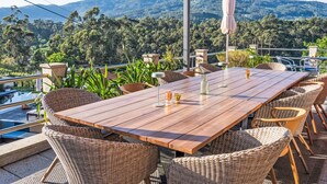 A wooden table and wicker chairs under a pergola with a mountain view.