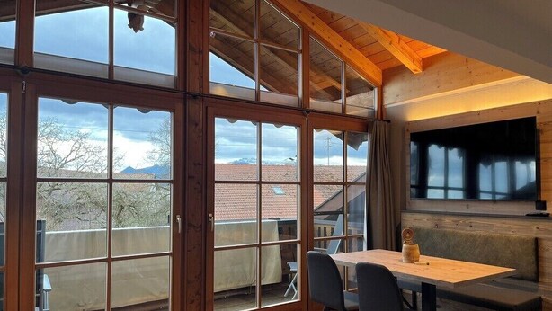 Ein Zimmer mit großen Fenstern, einer Holzbalkendecke, einem Tisch mit Stühlen und einer Bank.
