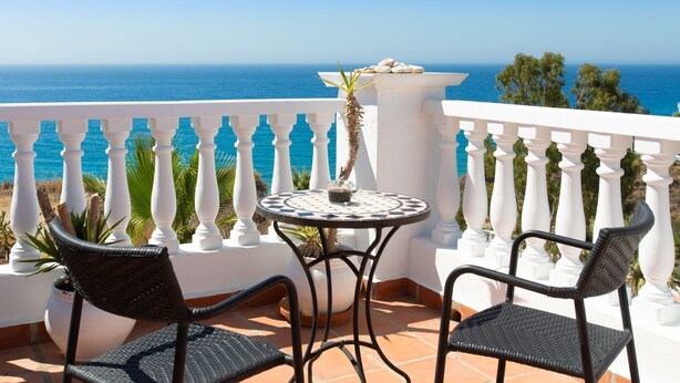 A balcony with a white balustrade, two wicker chairs, a round table with a patterned tablecloth, and a view of the sea.