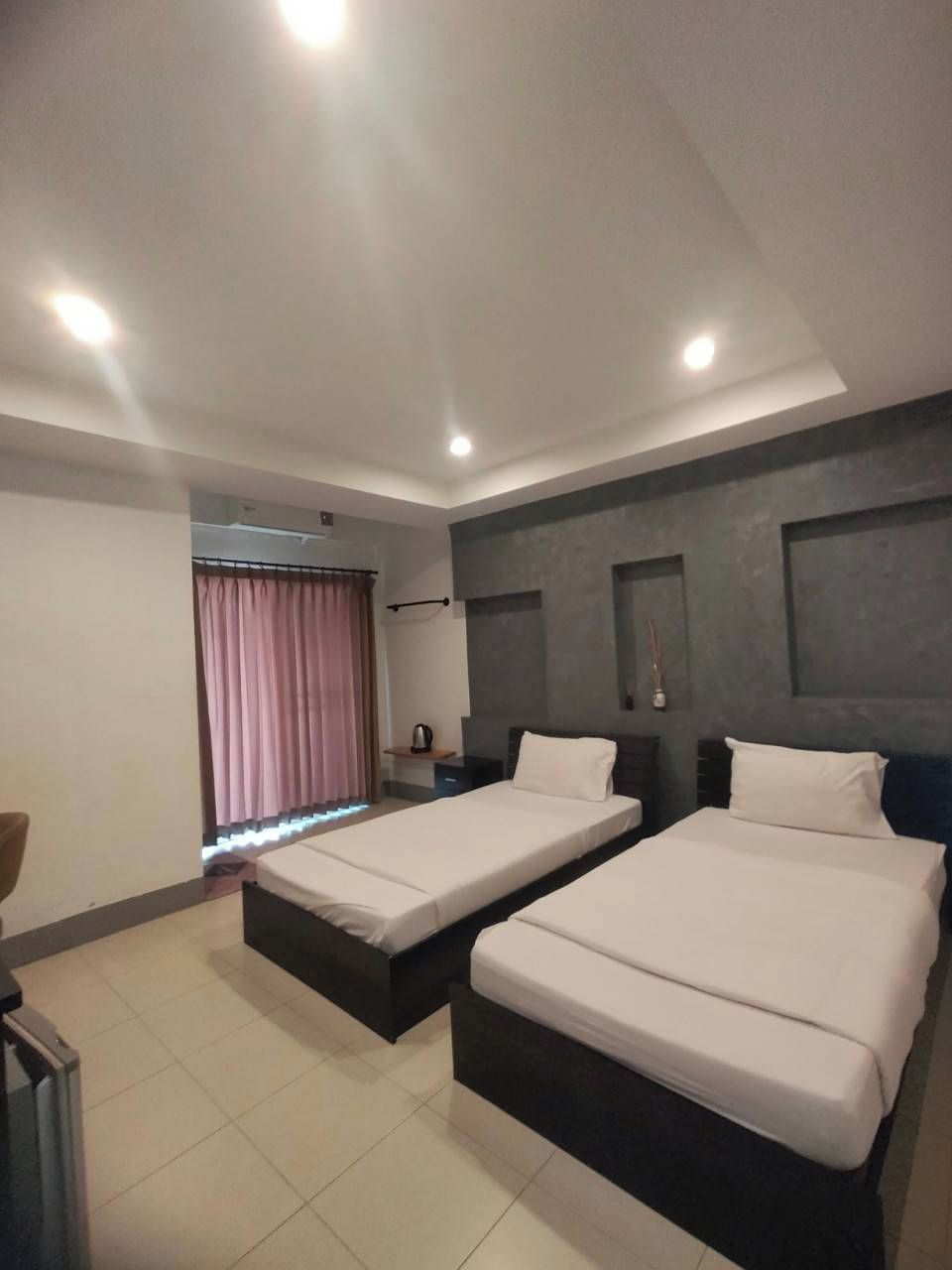Habitación de hotel moderna con dos camas individuales, un escritorio pequeño, una silla y un ventanal con cortinas.