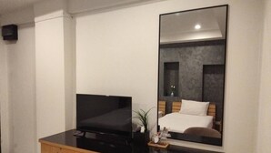 A hotel room with a bed, a desk with a television, a small refrigerator, and a large mirror. - Pilatus Apartment (Mueang Loei)