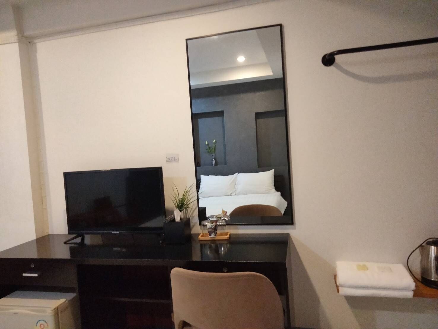 A hotel room with a flat-screen TV on a desk, a bed with white linens, a small potted plant, and a towel on the side.