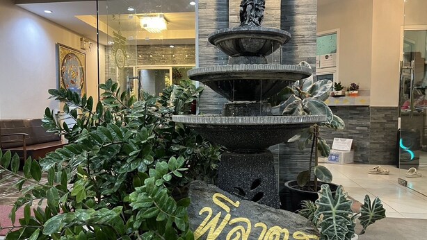 A lobby with a multi-tiered fountain, lush green plants, and a stone with text. - Pilatus Apartment (Mueang Loei)