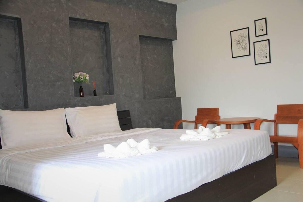 Cama bem arrumada com lençóis e toalhas brancas, uma cadeira de madeira e uma mesinha.