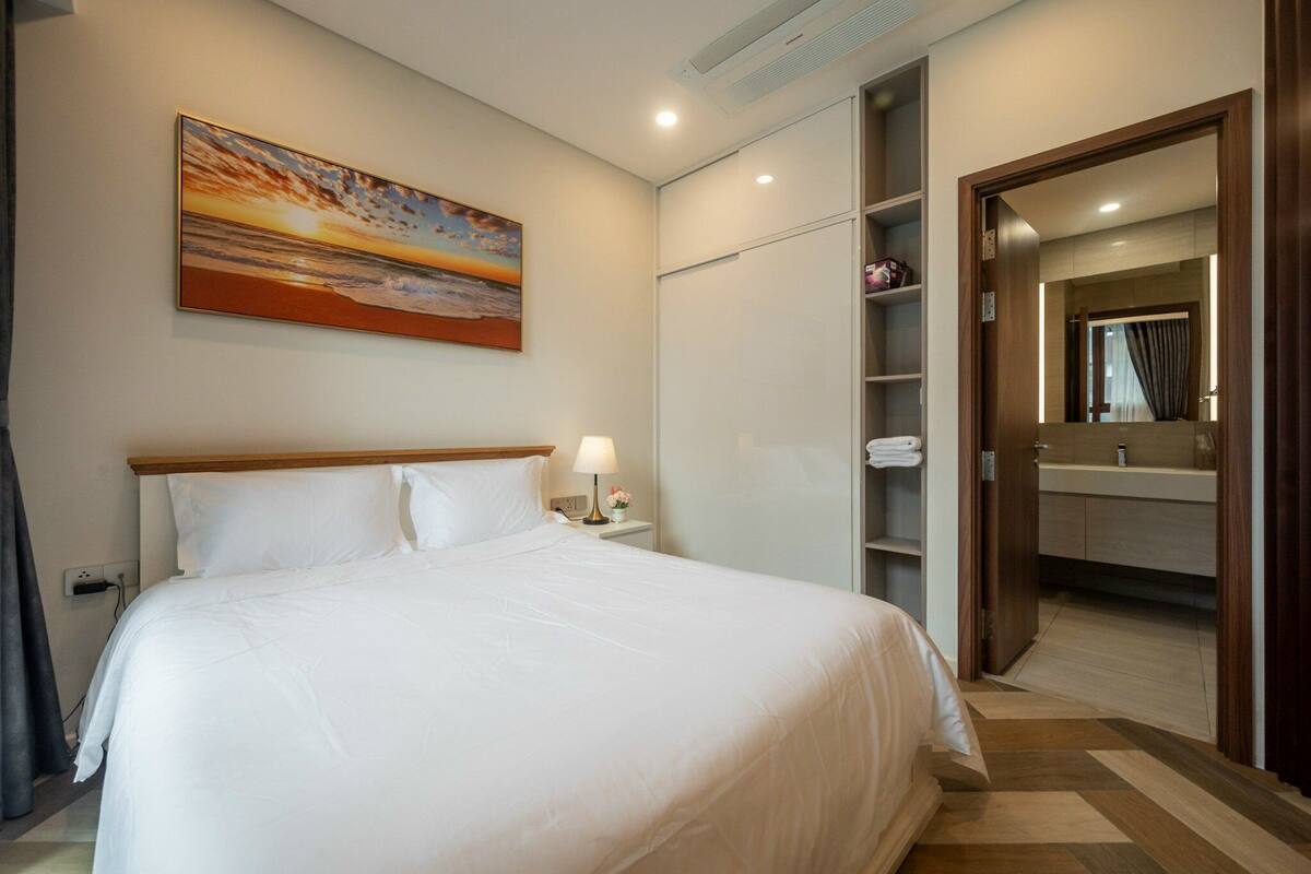 A neatly made bed with white linens, a bedside lamp, and a painting of a sunset on the wall.