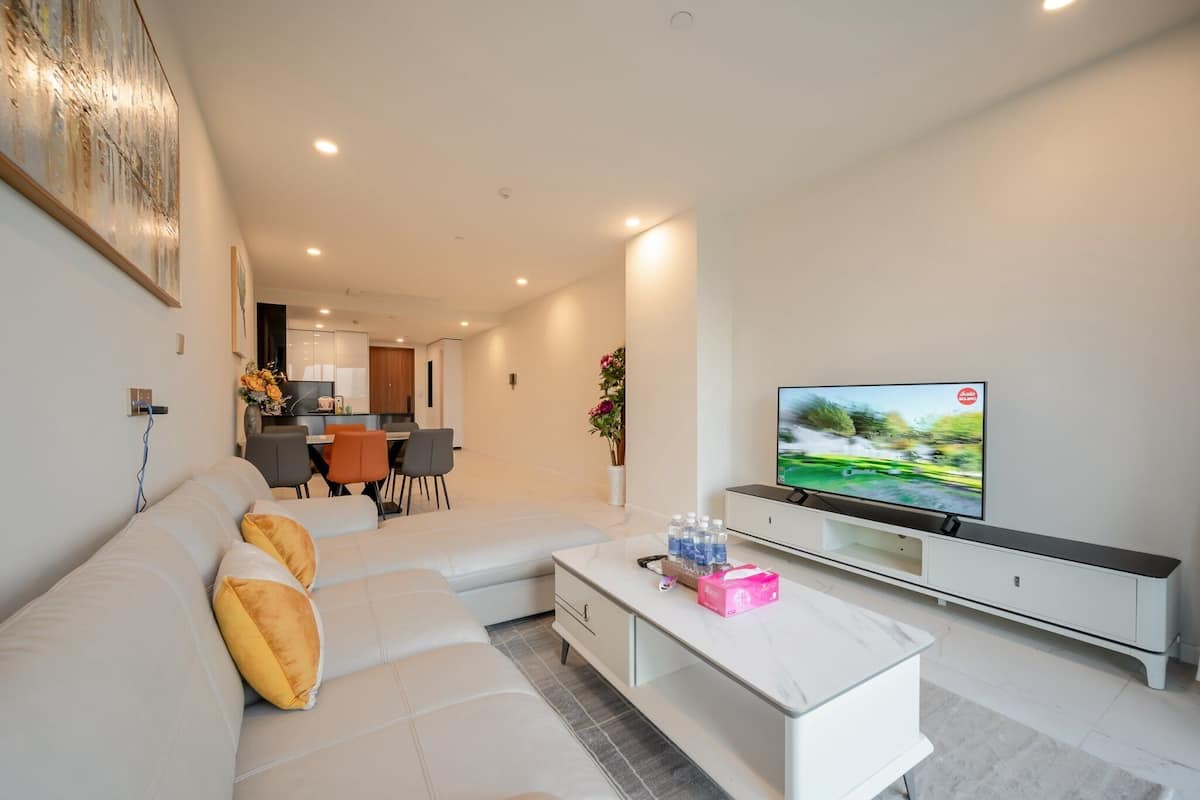 A modern living room with a white sofa, a coffee table, and a flat-screen TV. There is a dining area in the background with a table and chairs.