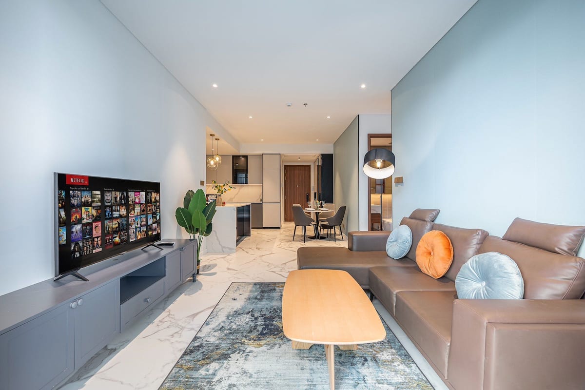 A modern living room with a flat-screen TV, a sofa, a coffee table, and a dining area in the background.