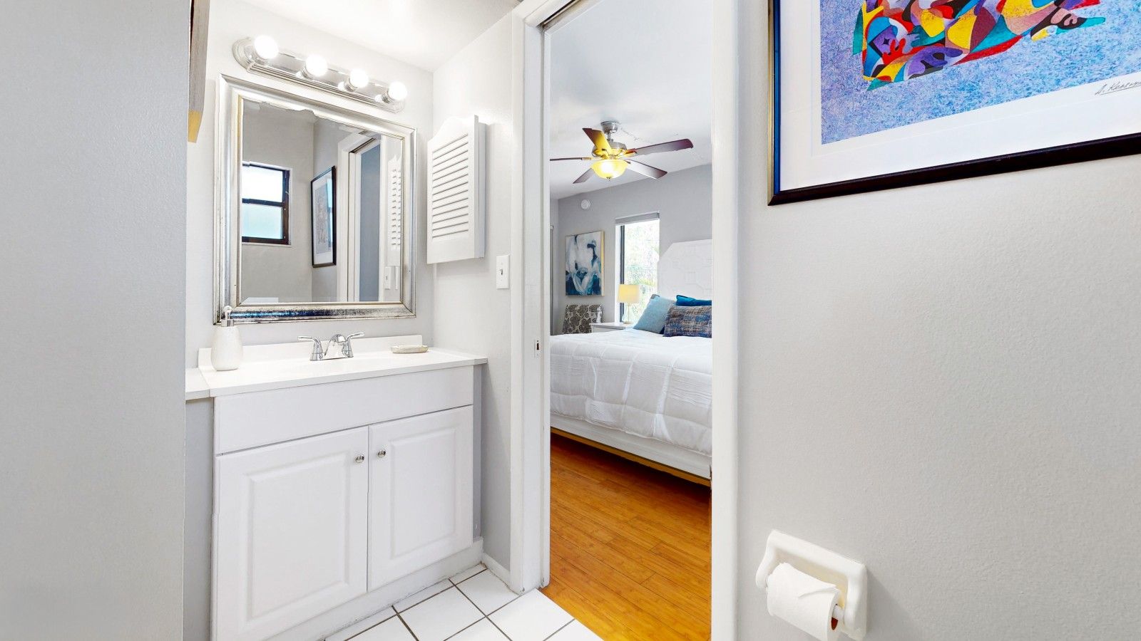 A bathroom with a white vanity, a mirror with lights, a ceiling fan, and a bed with a white duvet in the adjacent room.