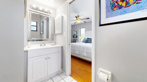 Une salle de bain avec un meuble-lavabo blanc, un miroir avec éclairage intégré, un ventilateur de plafond, et une chambre adjacente avec un lit recouvert d’une couette blanche.