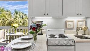 A kitchen with a white stove, sink, and cabinets, a dining table with plates, and a view of palm trees. - Beach Rd, 300ft to Beach Access & 500ft to Siesta Key Village, Studio (Siesta Key)