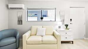 Ein modernes Wohnzimmer mit einem beigefarbenen Sofa, einem blauen Sessel, einem Fenster mit Blick auf Gebäude, einem weißen Beistelltisch mit Lampe und einer Klimaanlage.