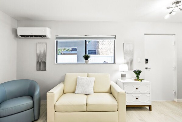 A modern living room with a beige sofa, a blue armchair, a window with a view of buildings, a white side table with a lamp, and an air conditioning unit. - Beach Rd, 300ft to Beach Access & 500ft to Siesta Key Village, 1bdr (Siesta Key)