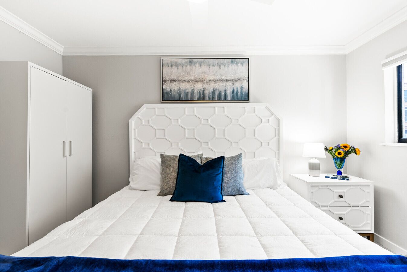 A bedroom with a white bed, a geometric headboard, a white wardrobe, and a bedside table with a vase of flowers.