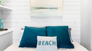 Une chambre à coucher avec un lit, des coussins turquoise, un coussin blanc orné du mot « PLAGE », une étagère avec un objet décoratif et un tableau accroché au mur.