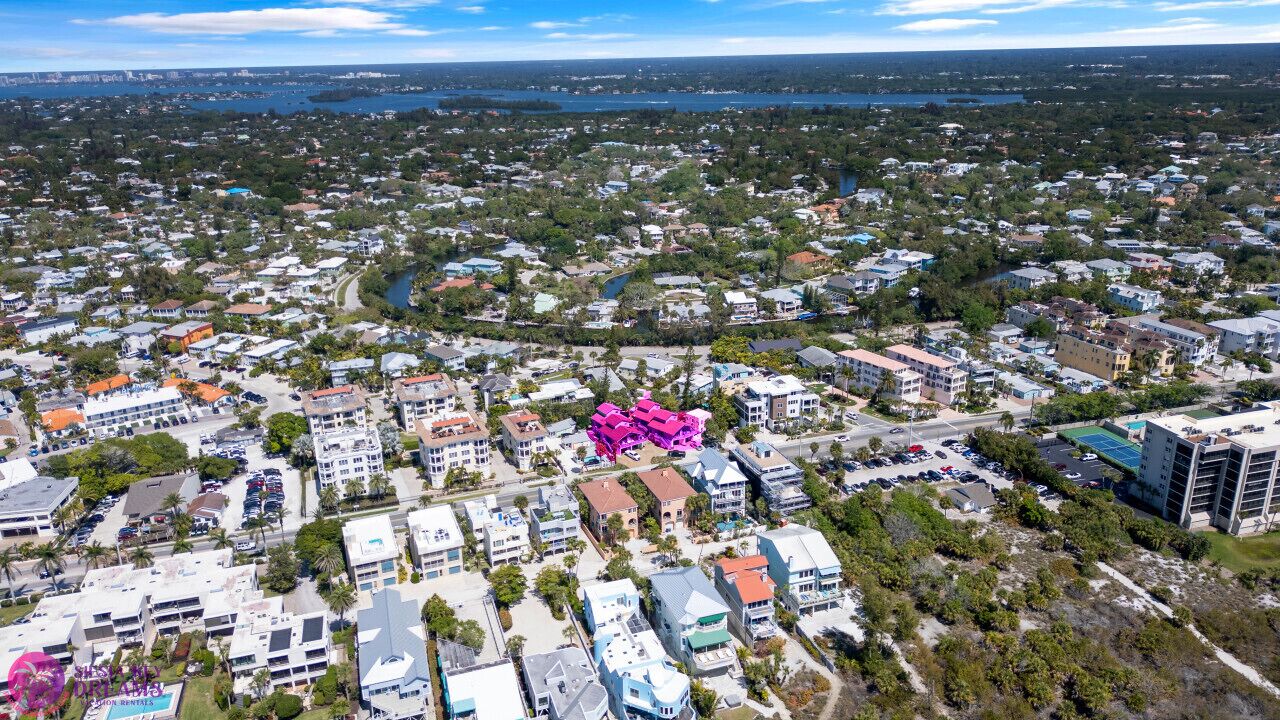 An aerial view of a residential area with a mix of single-family homes and apartment buildings, surrounded by greenery and a body of water in the distance.