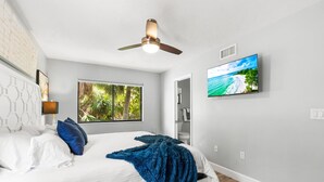 Une chambre à coucher avec un lit, un ventilateur de plafond, un téléviseur fixé au mur et une fenêtre donnant sur des arbres.
