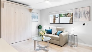 A modern living room with a beige sofa, glass coffee table, and two side tables. There is a ceiling fan, a large window with a view of greenery, and two framed artworks on the wall. - Beach Rd, 300ft to Beach Access & 500ft to Siesta Key Village, 1bdr (Siesta Key)